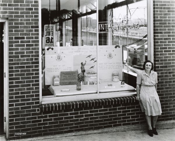 Mrs. Fey D. Murray(?) stands outside a storefront window display  promoting soybean oil for national defense during World War II. Exterior view of woman sitting on window sill of brick building. The window display has a sign that reads: "Farmers will produce the oil we lost has your farm enlisted?" The display was prepared Mrs. Murray(?), a DeMoines County Triple A farm field woman(?).