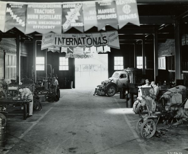 Mechanics work on tractors and a truck in the service shop of an International Harvester dealership. Advertising banners hang from the ceiling and in the background is a large open doorway.