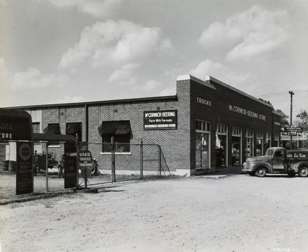 Exterior view of the storefront of an International Harvester dealership with a truck parked in front. On the left is a chain-link fence, with a sign in the shape of a man pointing that says: "LOOK / USED TRUCKS AND TRACTORS."