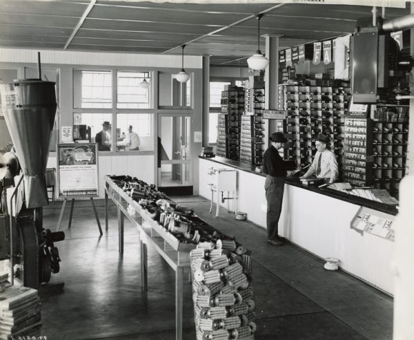 Two men at a parts counter in an International Harvester dealership. There is a water cooler, advertising poster, hammer mill and parts bins in the store. Two men are talking in an office in the background.