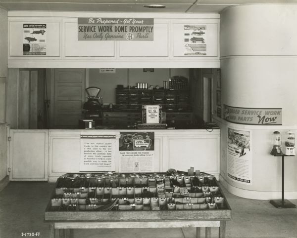 Interior view of the Kraft & Starr Company, an International Harvester dealership. There is a table of parts, gumball machines, and advertising posters for trucks and farm equipment. A sign above the counter reads: "Be Prepared-Get Your Service Work Done Promptly / Use Only Genuine IH Parts."