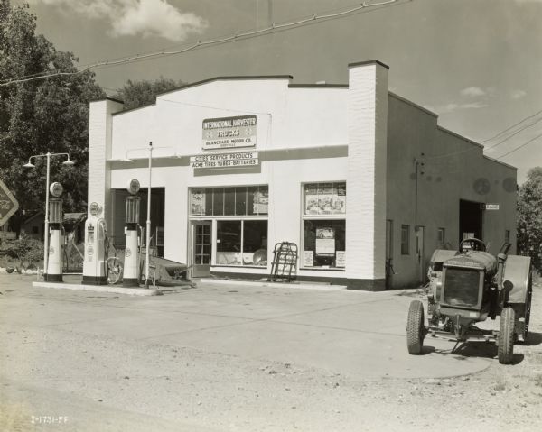 Exterior of Blanchard Motor Company service station. Blanchard Motor Company was an International truck dealership. A McCormick-Deering tractor and manure spreader are parked outside the building. "Cities Service" gas pumps stand in front of the building.