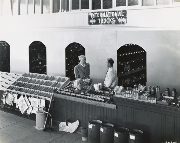 Two men stand behind a parts counter at the Arthur C. Day Implement Company, an International Harvester dealership. A sign above the counter reads: "International Trucks."