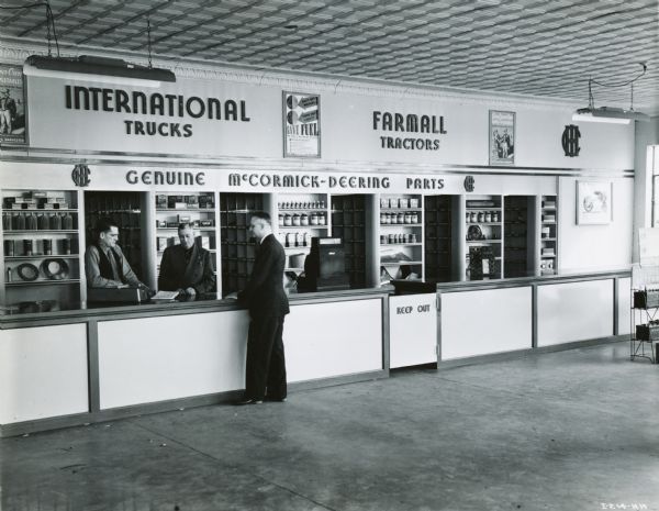 Three men stand at the parts counter of the Bush-Terry Implement Company, an International Harvester dealership. Signs advertising "International Trucks" and "Farmall Tractors" appear over the counter, and parts bins are in the background. According to the original caption, the store was operated by R.B. Bush and J.B. Terry.