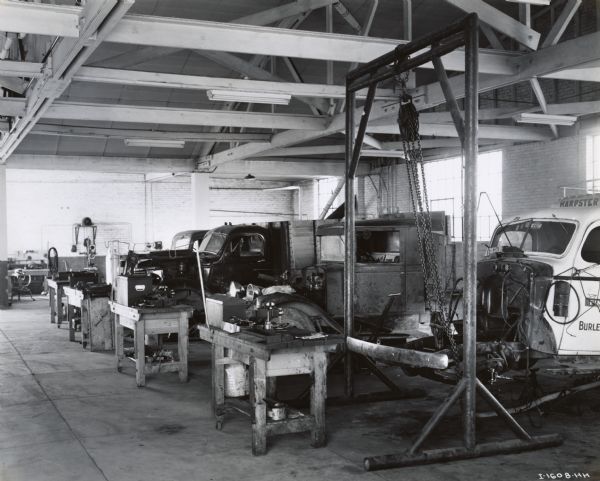 Four trucks in the shop or garage at Chisholm Brothers, an International Harvester dealership in Snake River Valley, Idaho. There are tables with tools and automotive parts on them. The front of a truck chassis is suspended from a pulley on the right.