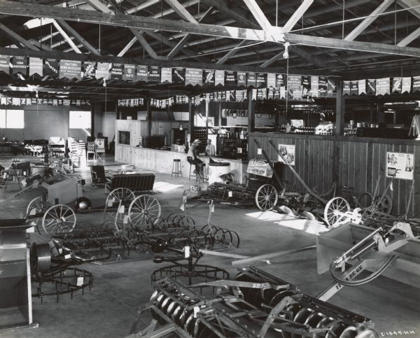 Showroom floor and service counter of the Brown Tractor Company, an International Harvester dealership. Two men are at the service counter. Advertising posters and banners are posted throughout the showroom. Farm implements on display include harrows, plows, a feed grinder and wagon truck. The dealership was operated by Burr Brown and A.E. Greiner.