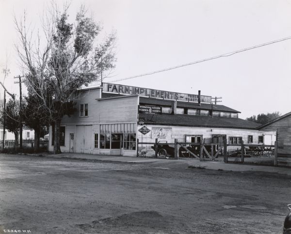 Exterior view from across road of the storefront of an International Harvester agricultural equipment dealership with a fenced-off side yard. Advertisements for "Chesterfield," "Goodyear" and "McCormick-Deering Service" are on the side of the building. A manure spreader is parked in the yard.
