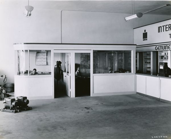 Three men working in offices at the Bush-Ellis Implement Company, an International Harvester dealership. On the right is a service counter, and on the left stationary engines are displayed on the floor.