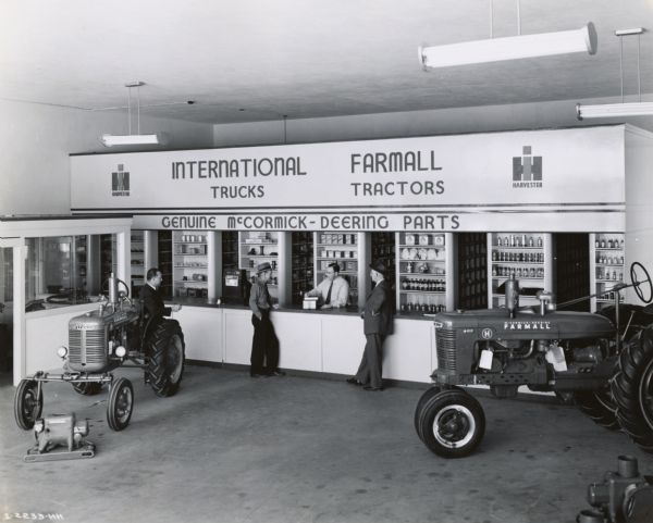 Men at the parts counter of the Bush-Ellis Implement Company, an International Harvester dealership. The man behind the parts counter is James F. Ellis, co-owner of the dealership. J.I. Chitwood, assistant manager of an International general line branch, is leaning on a Farmall A tractor at left. There is a Farmall H tractor also on display.