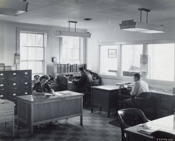 One woman and two men working at desks in an office at Industrial Equipment Company, an International Harvester dealership.