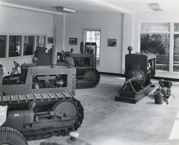 International crawler tractors (TracTracTors) and a power unit on display in a showroom at Industrial Equipment Company, an International Harvester dealership. A poster in the background reads, in part: "Back the Attack!"