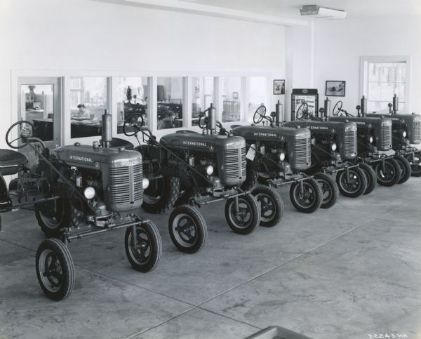 A line of Farmall A tractors on display in a showroom at the Industrial Equipment Company, an International Harvester dealership. Behind a set of windows on the back wall a woman is working in an office. A poster in the background reads, in part: "Back the Attack!"