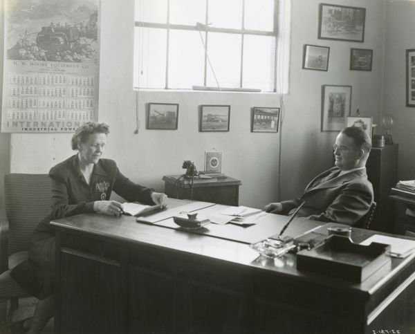 John C. Moore and Louise Strauss sitting at a desk at H.W. Moore Co., an International power equipment dealership. Mr. Moore was president of the company and Ms. Strauss was treasurer. A telephone, advertising calendar and framed photographs are in the background.