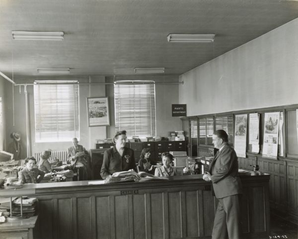 John C. Moore (right) standing at the cashier counter in the offices of H.W. Moore Equipment Company, an International Harvester power equipment dealership. A man and several women are working at desks in the background.