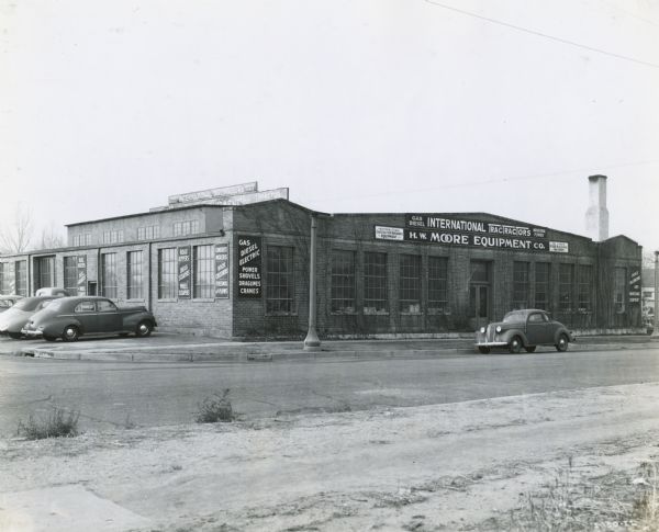 View from across road of the front of the H.W. Moore Equipment Company, an International Harvester power equipment dealership. Signs on the building read: "Gas Diesel International TracTracTors" and "Gas Diesel Electric Power Shovels Draglines, Cranes." Cars are parked next to the building.