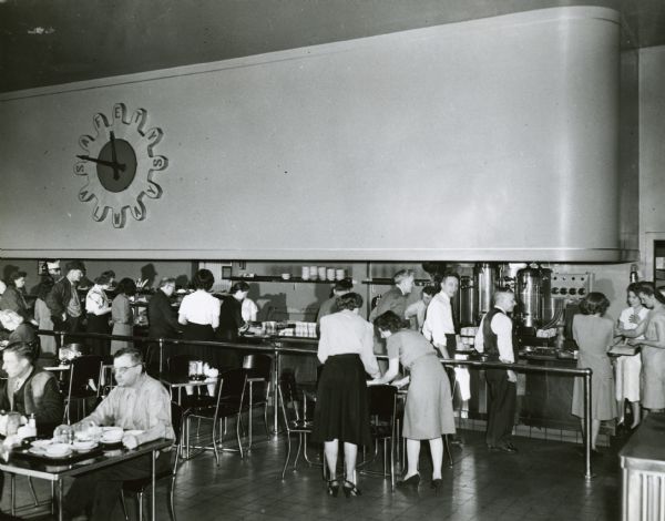 Factory workers sitting at tables and standing in line to get food at a service counter in the cafeteria of International Harvester's Tractor Works. The clock on the wall behind the counter reads: "Safety Always."