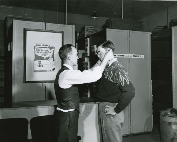 Two men standing in a factory office at International Harvester's Tractor Works. One man is fitting safety glasses on the other man. Behind them is a poster which reads: "Slim Chance says, let's put safety on the map in 1948! / Works Safety Committee."