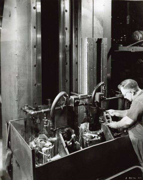 A factory worker wearing eyeglasses is holding a metal part while standing at a large machine in International Harvester's Tractor Works.