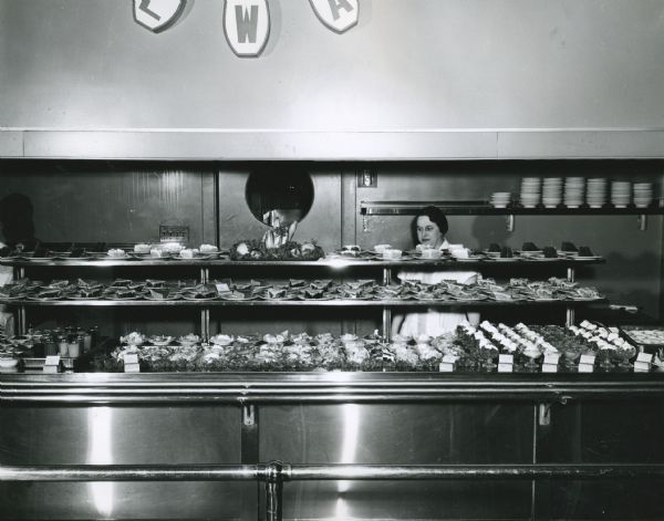 A woman standing behind a counter at the Tractor Works cafeteria. The shelves in front of her are filled with slices of pie on plates and ice cream in bowls.