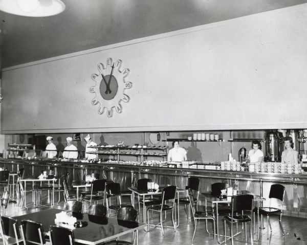 Three men and three woman standing behind a service counter at the cafeteria in International Harvester's Tractor Works (factory). The clock on the wall behind them reads, "Safety Always." The room is filled with tables and chairs.