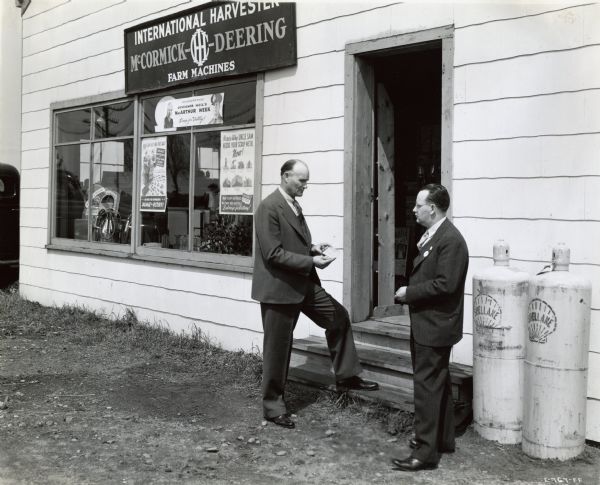 Two men involved in a scrap drive stand near the door of an International Harvester dealership. There are posters in the window. Next to the man on the right there are two white propane tanks which read, "Shellane."