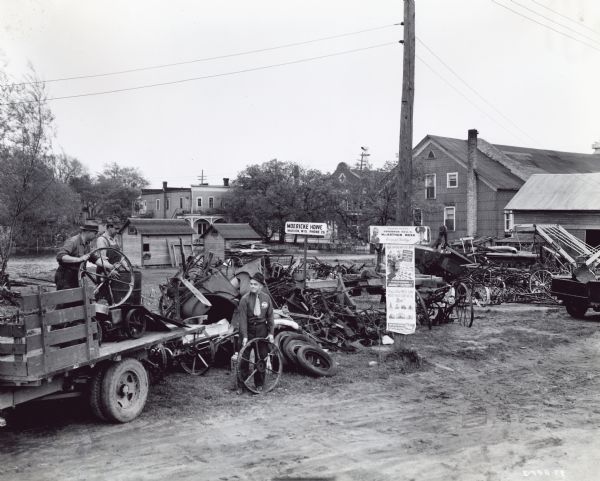 Three men loading a trailer near an International Harvester dealership during the "MacArthur Week" scrap drive. There is a large pile of scrap on the ground. A sign in the background says: "Moericke Howe, Marion, Wis. Phone 26."