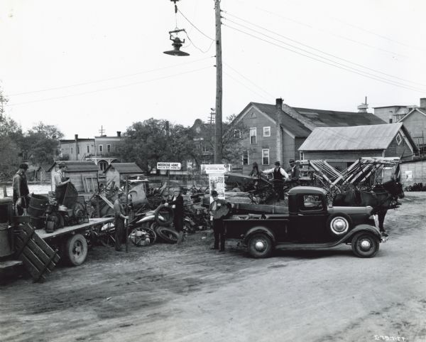 Men working on a "MacArthur Week" scrap drive near an International Harvester dealership. Two men are on the flatbed of a truck, and there is also a horse-drawn vehicle. A sign in the background reads: "Moericke Howe, Marion, Wis. Phone 26."
