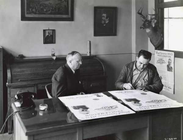 G.M. Ahlschwede, branch manager of Green Bay, watches R.P. Dunham, dealer of Green Bay, sign Governor Heil's pledge of cooperation, which reads, in part: "MacArthur Week Scrap Collection for Victory." A stuffed deer head is on the wall behind them.