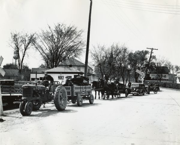 Men hauling wagon loads of scrap metal with a Farmall H tractor, horses, and an automobile. The men are participating in a scrap drive, possibly during Governer Heil's "MacArthur Week." There are houses, an American flag, and a water tower in the background.