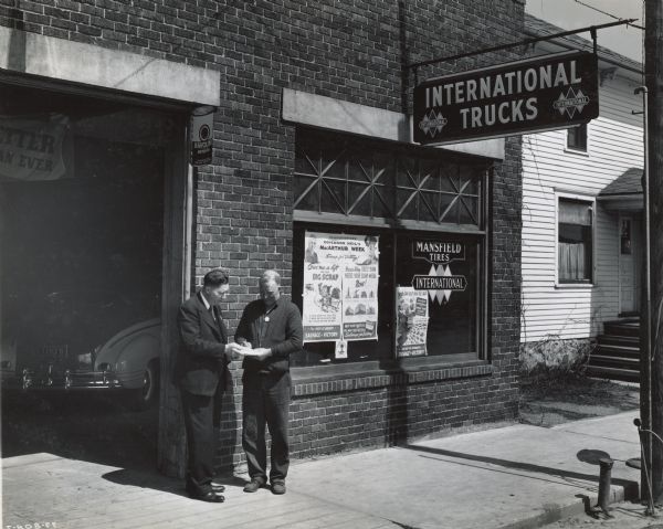 Blockman A.W. Olson and Lee Murphy outside the latter's International Harvester dealership. There is a sign for "International Trucks" above the sidewalk. There is a poster in the window for the: "MacArthur Week Scrap Collection for Victory." A parked car is in the background in the garage.