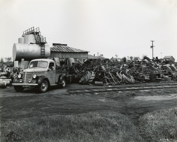 A man using an International truck to haul scrap metal for J.G. Larson & Sons, an International Harvester dealership. The man is dropping the metal off at a scrap pile near railroad tracks as part of a scrap drive effort.