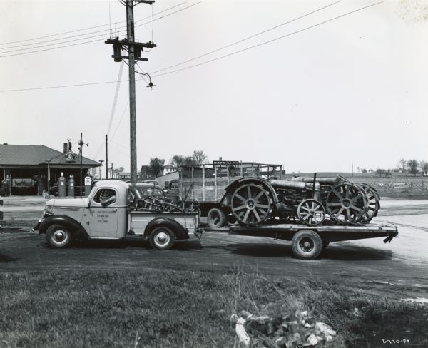 Man pulling a trailer with an International truck for J.G. Larson & Sons, an International Harvester dealership. The trailer is loaded with two old tractors, collected for a scrap drive. Gas pumps and a service station are in the background.