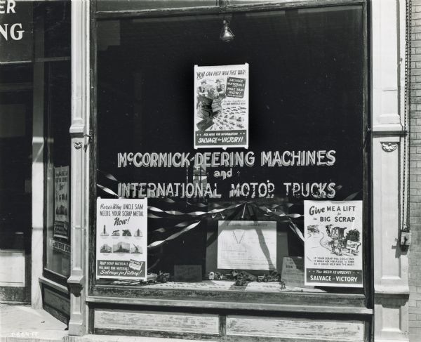 Close-up of a storefront window of the Burlington Farm Machinery Corporation, an International Harvester dealership. The window display includes posters promoting scrap drives.
