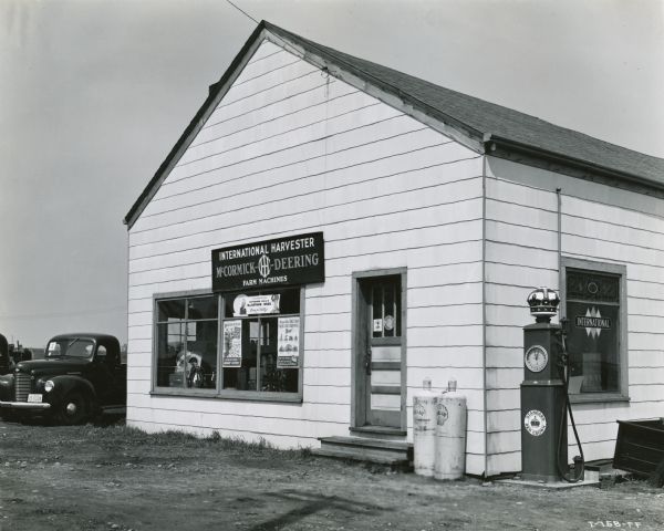 Exterior of the Will Implement Company, an International Harvester dealership. There are posters in the windows next to the front door, and on the right there are two white propane tanks which read, "Shellane," and also a Standard Red Crown gas pump.