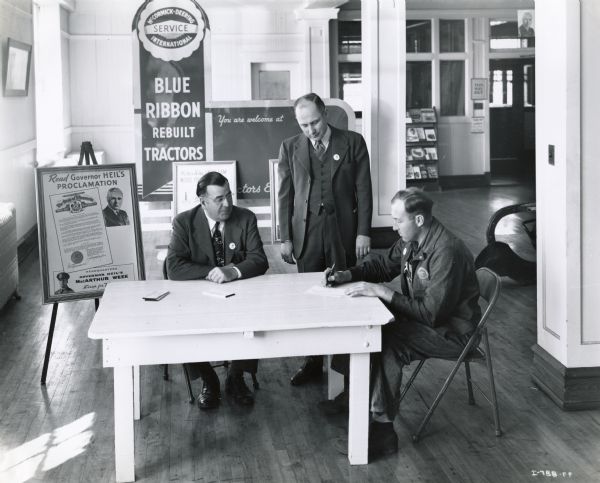 International Harvester dealer Chester E. Seif signs a pledge of cooperation with Governor Heil's "MacArthur Week" scrap drive. H.J. Brosnahan (left), manager of the Eau Claire branch, and blockman W.H. Tudor look on. Posters are displayed throughout the dealership showroom and a literature rack is just visible in the background. One poster reads: "Blue Ribbon Rebuilt Tractors", and another poster near the table reads: "MacArthur Week Scrap for Victory."