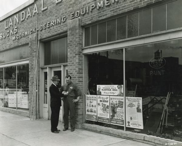 Milwaukee blockman Dale Thomas pins a "MacArthur Week" button on Charles Landaal, operator of Landaal Brothers, an International Harvester dealership. The storefront windows include scrap drive posters, toy wagons, toy tractors, gardening tools and a Mautz Paint sign.