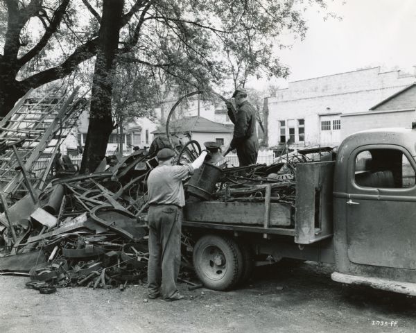 Two men unloading scrap metal from a truck near an International Harvester dealership, possibly Landaal Brothers of Waupun. One man is standing on the truck, and the other is standing on the ground. An industrial building and houses are in the background.