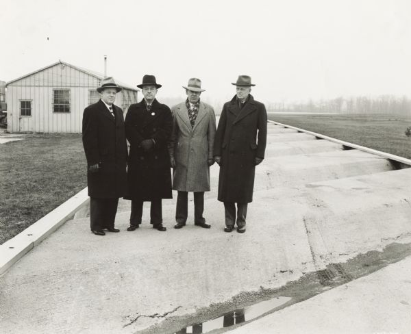 Four men in hats and overcoats standing on a deeply ridged concrete track on the proving grounds at International Harvester's Ft. Wayne Works. An original handwritten caption identifies the men as (left to right): L.W. Pierson(?); R.C. Nourse - Canadian Bell; H. Reinoehls(?) - Chief Engineer Ft. Wayne Works; T.C. Smith - Engineer AT&T New York.