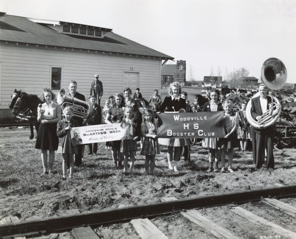 A group of children, including members of the Woodville High School Booster Club, stand near a pile of scrap metal. Some of the children are holding musical instruments. Others hold two banners, one of which reads: "Headquarters, Governor Heil's MacArthur Week, Scrap for Victory!" Railroad tracks are in the foreground.