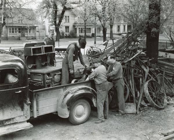 Three men unload scrap metal from a truck in the yard of Landaal Brothers, an International Harvester dealership. Dealer Charles Landaal is standing at right. There is a residential street with houses in the background.