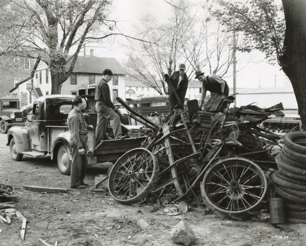 Farmers unload scrap metal from a truck in the yard of Landaal Brothers, an International Harvester dealership. Dealer Charles Landaal is standing at left. Houses are in the background.