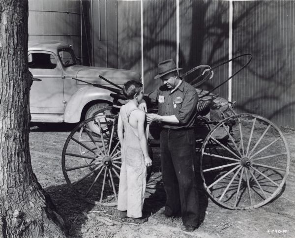 International Harvester dealer Albert Karpernich(?), Jr. pins a "MacArthur Week" button on a boy scrap collector. According to the original caption, Karpernich(?) was the Assistant County Chairman and Township Chairman for the scrap drive. A wagon, automobile, barn and silo are in the background.