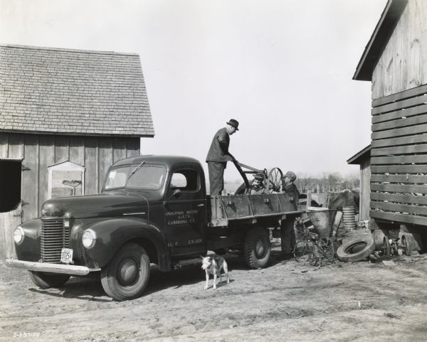 Men from Anderson Motor Sales, an International Harvester dealership, collect a load of metal scrap form the farm of Frank Cook. The men are loading the metal into the back of an International truck. The truck is parked among farm buildings and a small dog is wandering nearby.