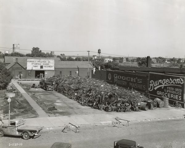 Elevated view of a large metal scrap pile in an empty lot at the Kronquest Implement Company, an International Harvester dealership. Billboards next to the lot advertise "Gooch's Poultry Mashes" and "Burgeson's Groceries."