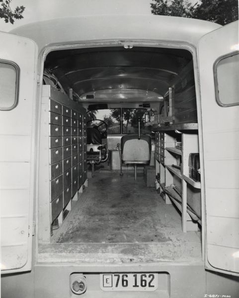 View of the interior of an International Metro truck through open rear doors. Inside the truck are a set of drawers on the left, and on the right is a counter with shelves for tools. A vise is attached to the counter. A driver and passenger seat are in the front of the truck.