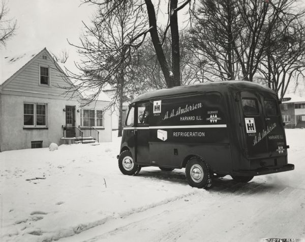Man inside an A.A. Anderson refrigeration truck parked outside a house in the snow. A.A. Anderson was an International Harvester dealership. The truck is a KB-3 with Metro body.