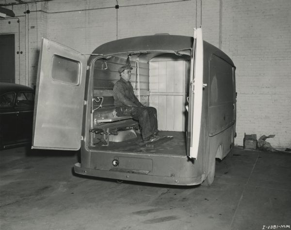 View through two open rear doors of a man seated on a bench in an unmarked Internatioanl Metro truck inside what appears to be a garage.