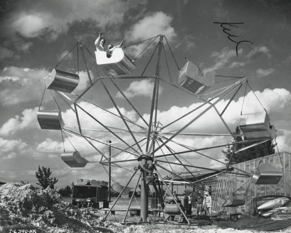 Two men aboard the Comet carnival ride, which is powered by International U-2 power units. Three men are standing on the ground near an industrial building.