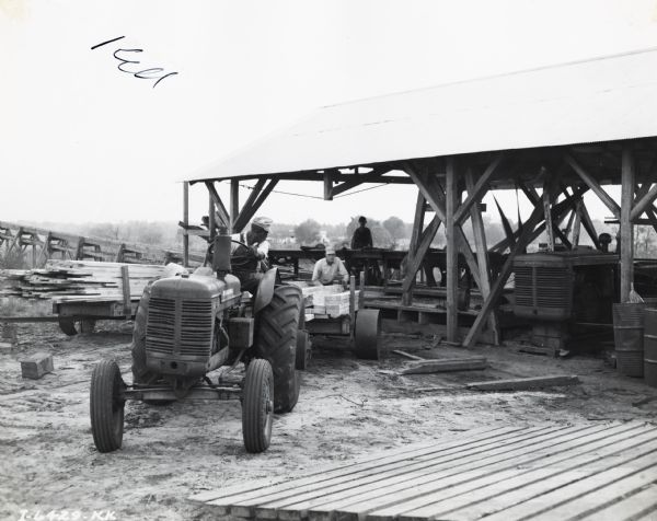 Man operates an International I-4 tractor while another man loads lumber onto the trailer at the Duke Lumber Company. Another man is standing under an open-sided roofed shed behind them.