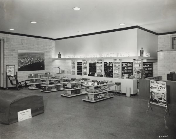 Interior view of International Harvester's Seattle branch house. In the foreground are shelves displaying products, and in the background is a service counter. An enlarged photograph or mural is displayed on the far back wall and a sign reads: "International Motor Trucks." A poster advertising the "Harvest of Stars" program on NBC is displayed at right.